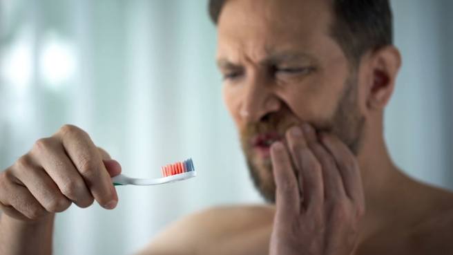 Man with bloody toothbrush in one hand and holding his cheek in pain with the other