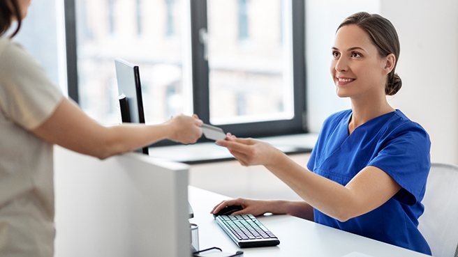 a woman wearing blue scrubs sitting at a desk accepting a credit card being handed to her