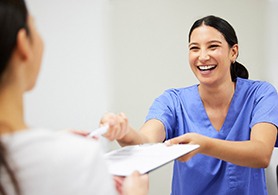 a woman wearing scrubs handing a patient forms to fill out on a clipboard