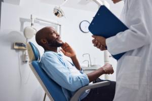 a man in a dental chair holding his mouth and speaking to a dentist with a clipboard